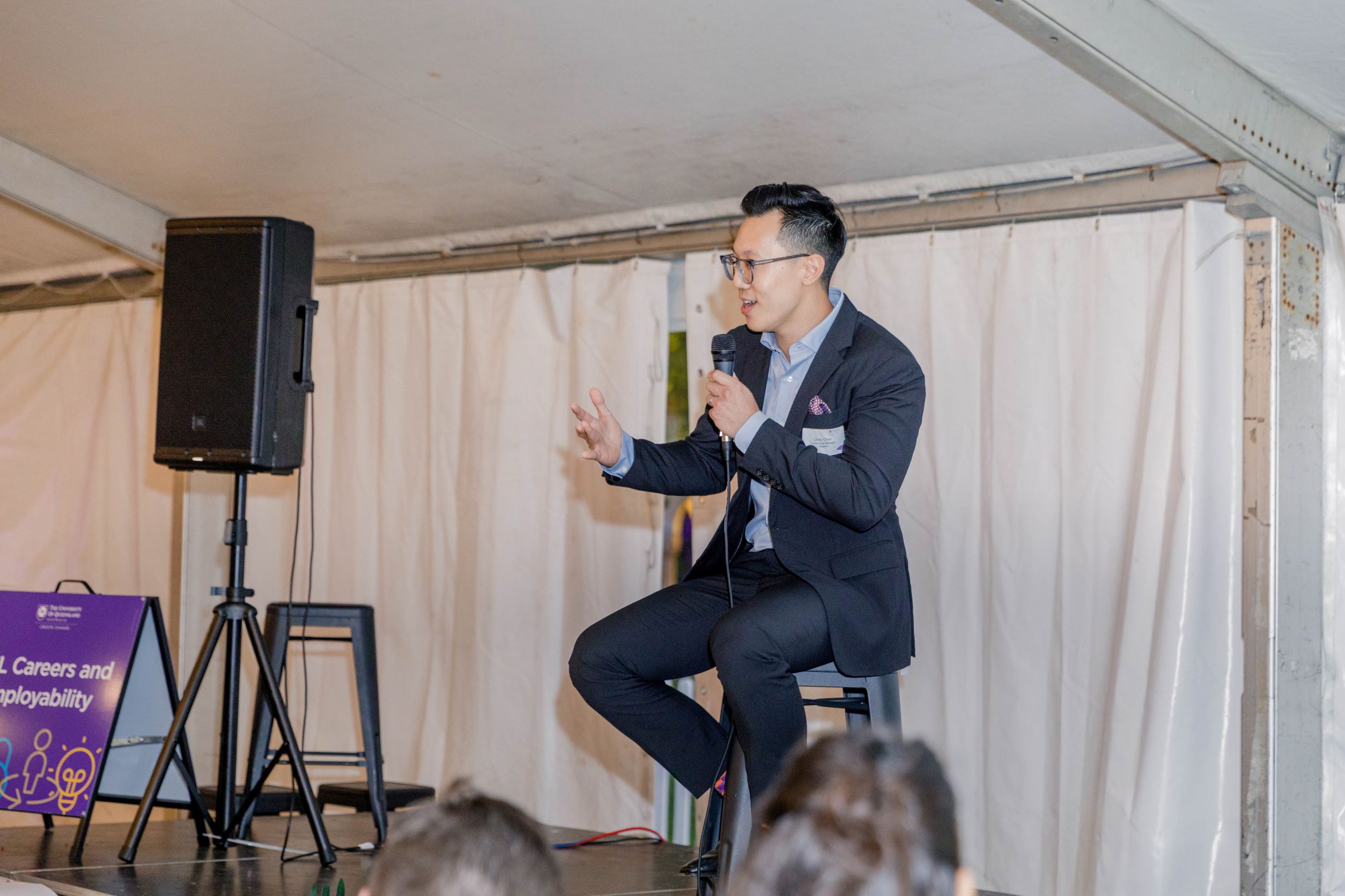 Chris Chan seated on a stool on a stage talking to an audience seated in front of him