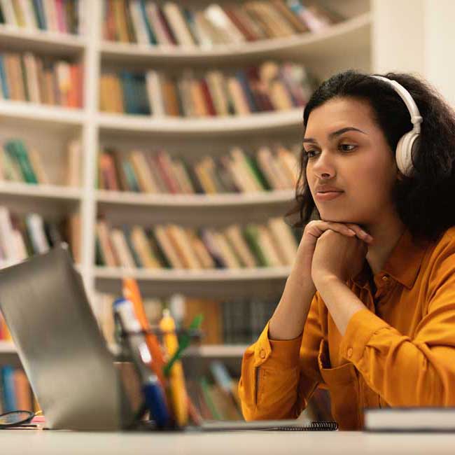 young woman studying at laptop in library with headphones on